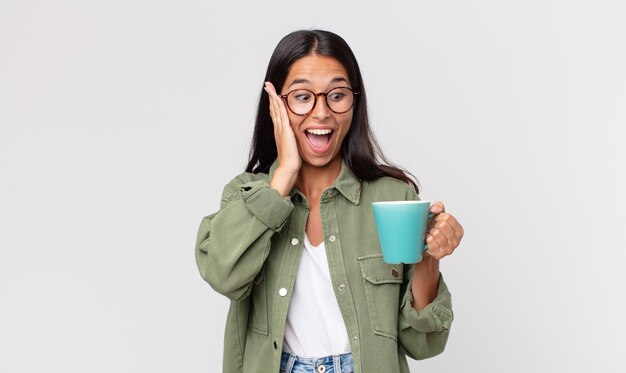 Jeune femme hispanique se sentant heureuse, excitée et surprise et tenant une tasse de café