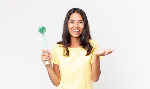 Jeune femme hispanique se sentant heureuse, surprise de réaliser une solution ou une idée et tenant une brosse à vaisselle