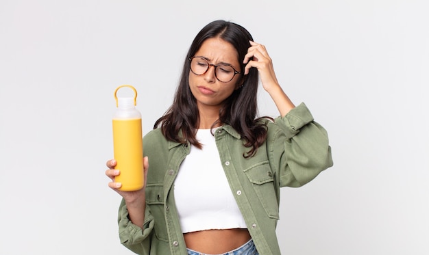 Jeune femme hispanique se sentant perplexe et confuse, se grattant la tête et tenant un thermos à café