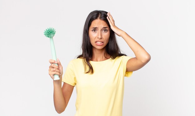 Jeune femme hispanique se sentant stressée, anxieuse ou effrayée, les mains sur la tête et tenant une brosse à vaisselle