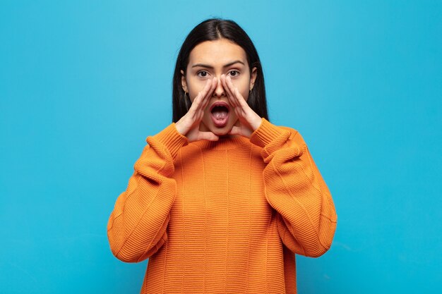 Jeune femme hispanique se sentir heureuse, excitée et positive, donnant un grand cri avec les mains à côté de la bouche, appelant