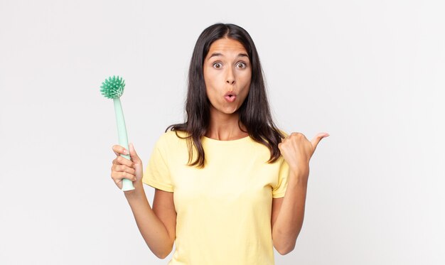 Jeune femme hispanique semblant étonnée d'incrédulité et tenant une brosse à vaisselle