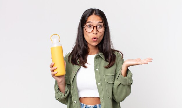 Jeune femme hispanique semblant surprise et choquée, avec la mâchoire tombée tenant un objet et tenant un thermos de café