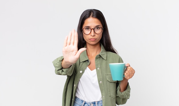 Jeune femme hispanique à la sérieuse montrant la paume ouverte faisant un geste d'arrêt et tenant une tasse de café