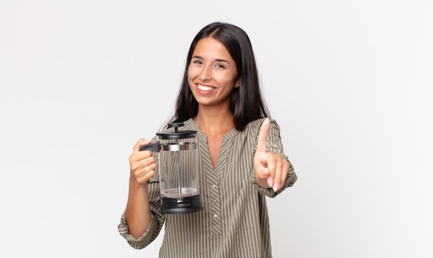 Jeune femme hispanique souriant fièrement et en toute confiance faisant le numéro un et tenant une cafetière manuelle