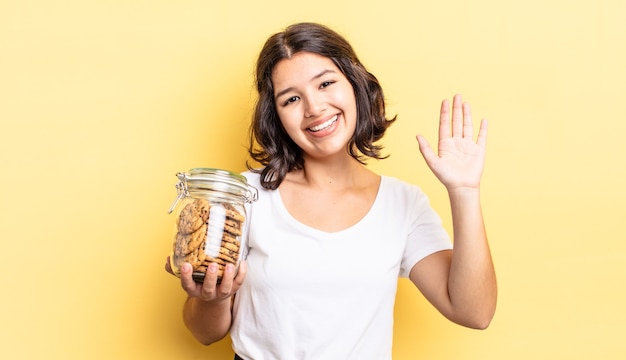 Jeune femme hispanique souriant joyeusement, agitant la main, vous accueillant et vous saluant. concept de bouteille de biscuits