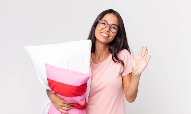 Jeune Femme Hispanique Souriant Joyeusement, Agitant La Main, Vous Accueillant Et Vous Saluant En Pyjama Et Tenant Un Oreiller