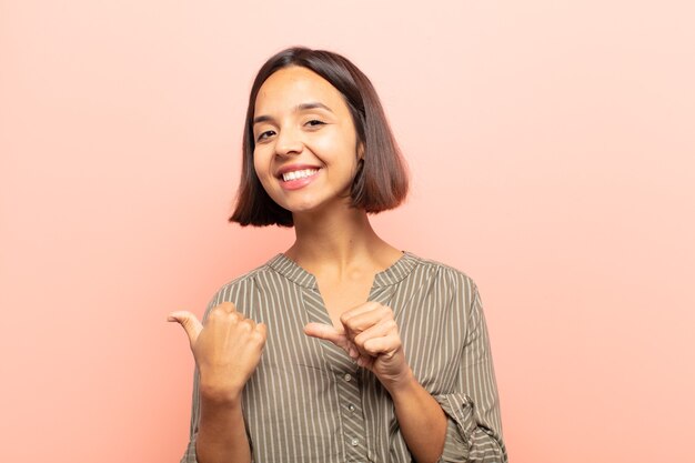 Jeune femme hispanique souriant joyeusement et pointant avec désinvolture pour copier l'espace sur le côté, se sentir heureux et satisfait