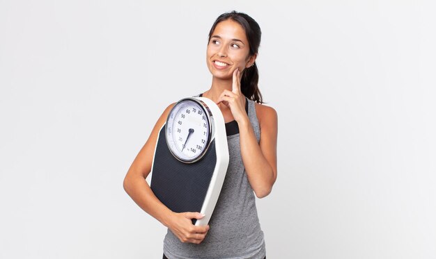 Jeune femme hispanique souriant joyeusement et rêvassant ou doutant et tenant un concept de régime d'échelle de poids