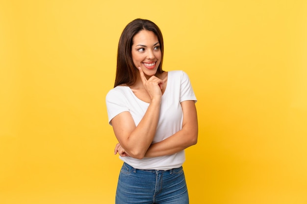 Jeune femme hispanique souriant joyeusement et rêvassant ou doutant