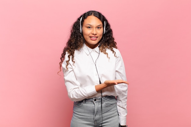 Jeune femme hispanique souriant joyeusement, se sentant heureuse et montrant un concept dans l'espace de copie avec la paume de la main. concept de télévendeur