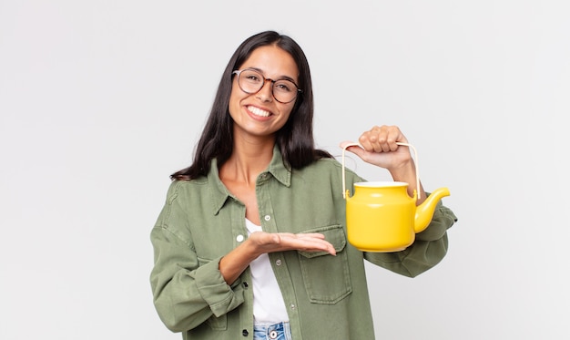 Jeune Femme Hispanique Souriant Joyeusement, Se Sentant Heureuse Et Montrant Un Concept Et Tenant Une Théière