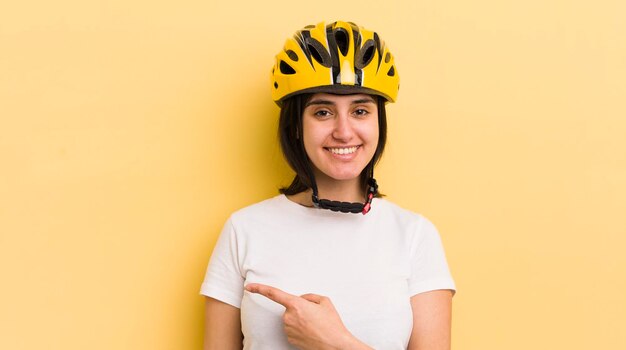 Jeune femme hispanique souriant joyeusement se sentant heureuse et pointant vers le concept de casque de vélo latéral