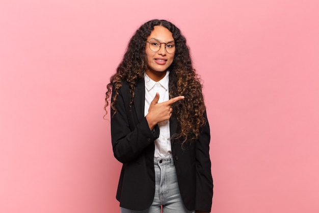 Jeune femme hispanique souriant joyeusement, se sentant heureuse et pointant vers le côté et vers le haut