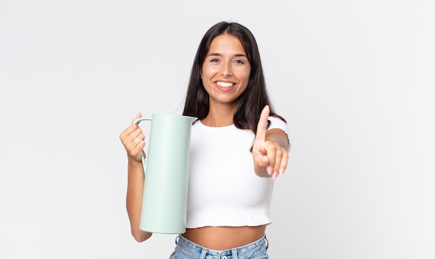 Jeune femme hispanique souriante fièrement et avec confiance faisant le numéro un et tenant un thermos de café