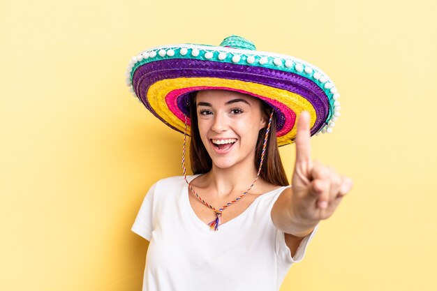 Jeune femme hispanique souriante fièrement et en toute confiance faisant numéro un. concept de chapeau mexicain