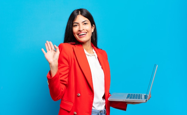 Jeune femme hispanique souriante joyeusement et joyeusement, agitant la main, vous accueillant et vous saluant, ou vous disant au revoir. concept d'ordinateur portable