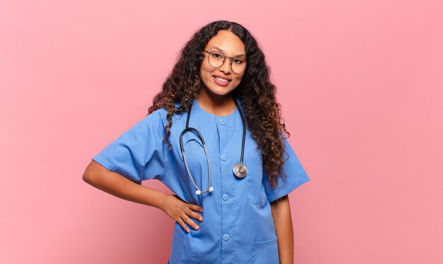 Jeune femme hispanique souriante joyeusement avec une main sur la hanche et une attitude confiante, positive, fière et amicale. concept d'infirmière