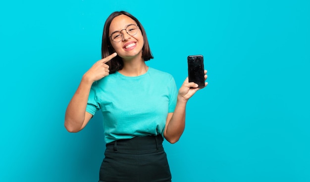 Jeune femme hispanique souriante pointant avec confiance vers son propre large sourire, attitude positive, détendue et satisfaite