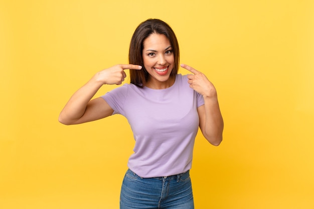 Jeune femme hispanique souriante pointant avec confiance vers son propre large sourire