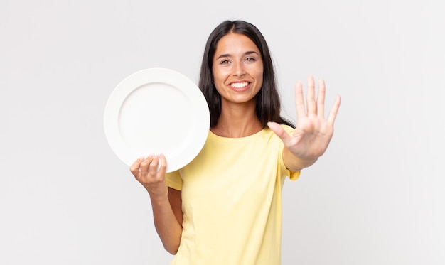 Jeune femme hispanique souriante et semblant amicale, montrant le numéro cinq et tenant une assiette vide