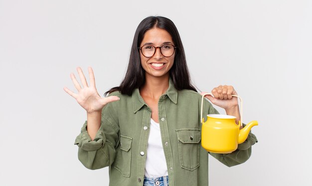 Jeune femme hispanique souriante et semblant amicale, montrant le numéro cinq et tenant une théière