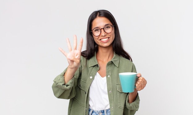 Jeune femme hispanique souriante et semblant amicale, montrant le numéro quatre et tenant une tasse à café