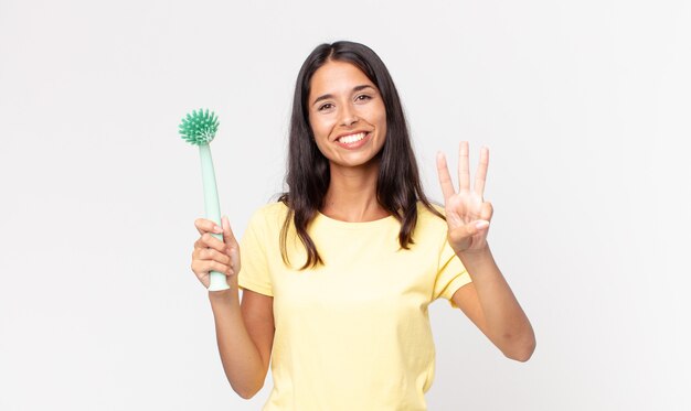 Jeune femme hispanique souriante et semblant amicale, montrant le numéro trois et tenant une brosse à vaisselle