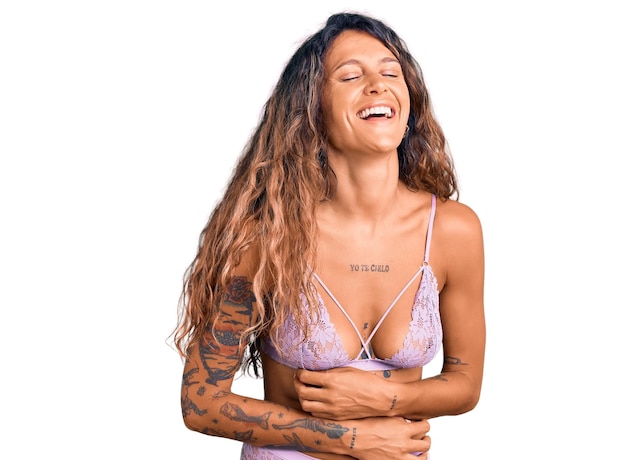 Jeune femme hispanique avec tatouage portant de la lingerie souriant et riant fort parce que drôle de blague folle avec les mains sur le corps