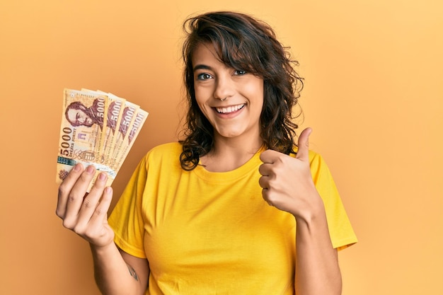 Jeune femme hispanique tenant 5000 billets en forint hongrois souriant pouce heureux et positif faisant un excellent signe d'approbation