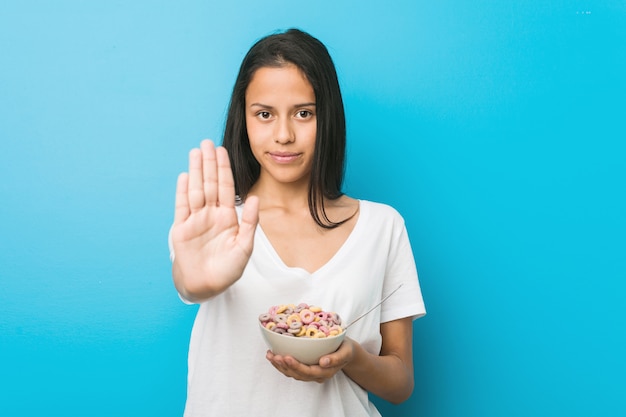 Photo jeune femme hispanique tenant un bol de céréales au sucre se tenant debout avec la main tendue montrant le panneau d'arrêt, vous empêchant.