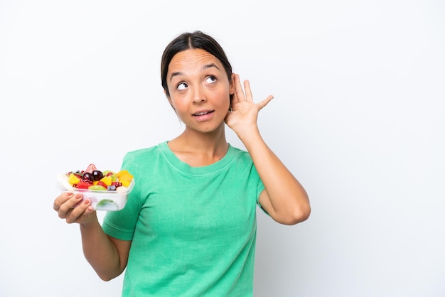 Jeune femme hispanique tenant un bol de fruits isolé sur fond blanc écoutant quelque chose en mettant la main sur l'oreille