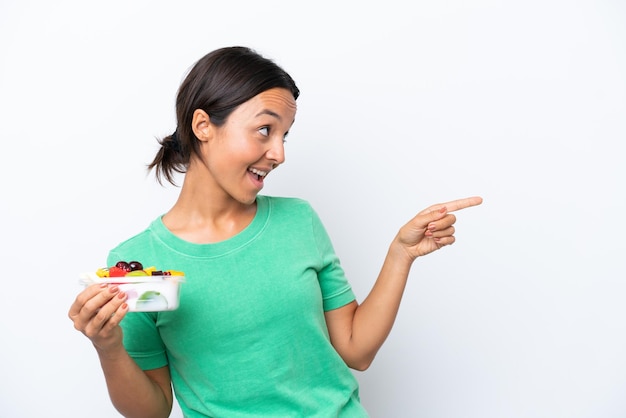 Jeune femme hispanique tenant un bol de fruits isolé sur fond blanc pointant le doigt sur le côté et présentant un produit