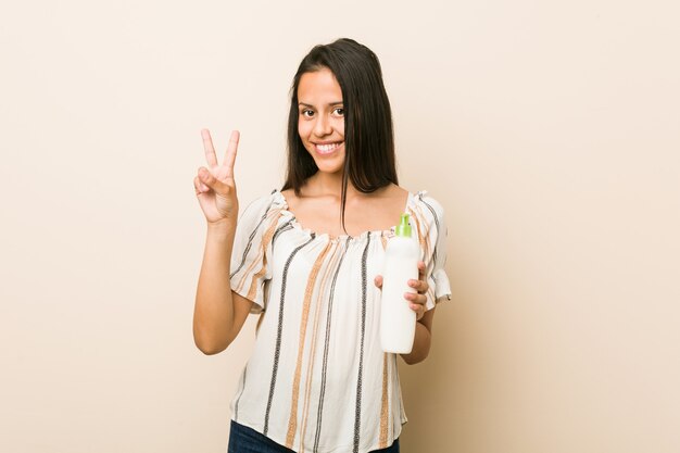 Jeune femme hispanique tenant une bouteille de crème numéro deux avec les doigts.