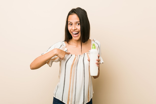 Jeune femme hispanique tenant une bouteille de crème surprise se montrant du doigt, souriant largement.