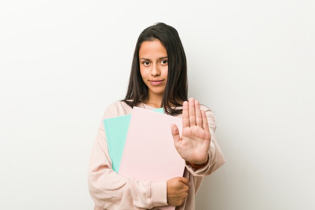 Jeune femme hispanique tenant des cahiers debout avec la main tendue montrant un panneau d'arrêt vous empêchant.