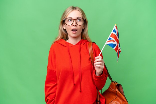 Jeune femme hispanique tenant un drapeau du Royaume-Uni sur fond isolé levant les yeux et avec une expression surprise