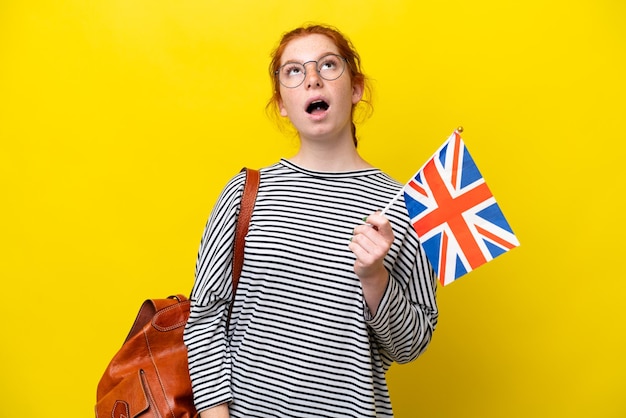 Jeune femme hispanique tenant un drapeau du Royaume-Uni isolé sur fond jaune en levant et avec une expression surprise