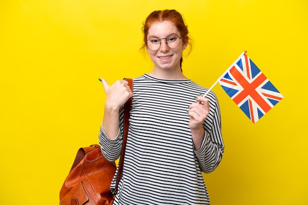 Jeune femme hispanique tenant un drapeau du Royaume-Uni isolé sur fond jaune pointant vers le côté pour présenter un produit