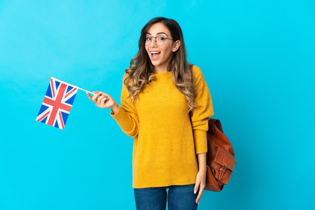 Jeune femme hispanique tenant un drapeau du Royaume-Uni isolé sur un mur bleu avec une expression faciale surprise