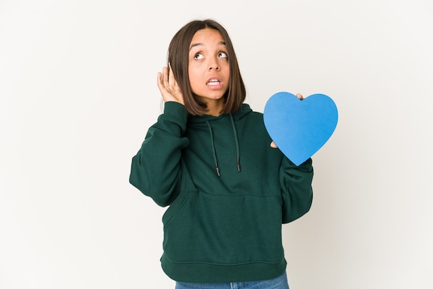 Jeune femme hispanique tenant une forme de coeur essayant d'écouter un commérage.