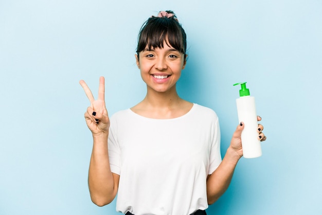 Jeune femme hispanique tenant une lotion pour le corps isolée sur fond bleu montrant le numéro deux avec les doigts