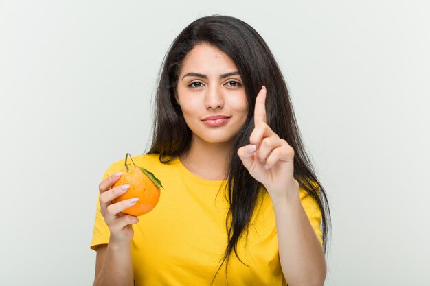 Jeune femme hispanique tenant une orange montrant le numéro un avec le doigt.