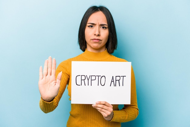 Jeune femme hispanique tenant une pancarte d'art crypto isolée sur fond bleu debout avec la main tendue montrant un panneau d'arrêt vous empêchant