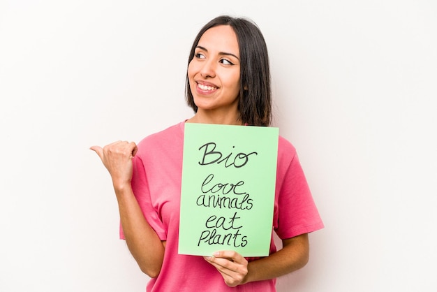 Jeune femme hispanique tenant une pancarte bio isolée sur fond blanc pointe avec le pouce en riant et sans souci