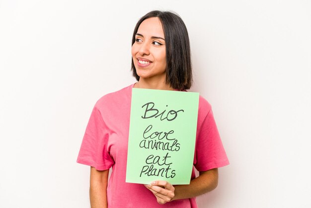 Jeune femme hispanique tenant une pancarte bio isolée sur fond blanc regarde de côté souriant gai et agréable