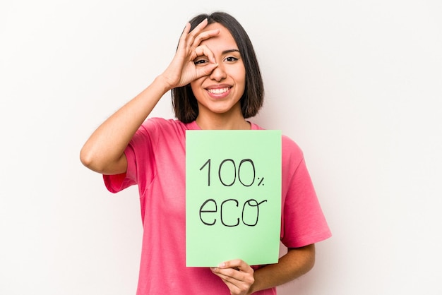 Jeune femme hispanique tenant une pancarte écologique 100 isolée sur fond blanc excité en gardant le geste ok sur les yeux