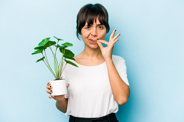 Jeune femme hispanique tenant une plante isolée sur fond bleu avec les doigts sur les lèvres en gardant un secret