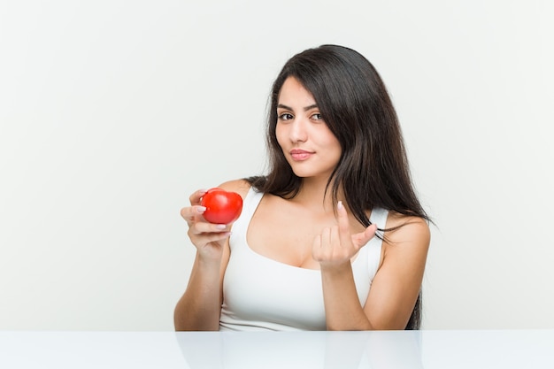 Jeune femme hispanique tenant une tomate pointant du doigt vers vous comme si vous vous invitiez à vous rapprocher.