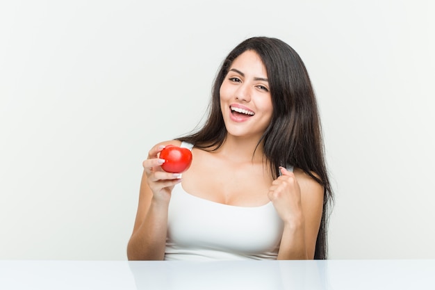 Jeune femme hispanique tenant une tomate surprise se montrant du doigt, souriant largement.